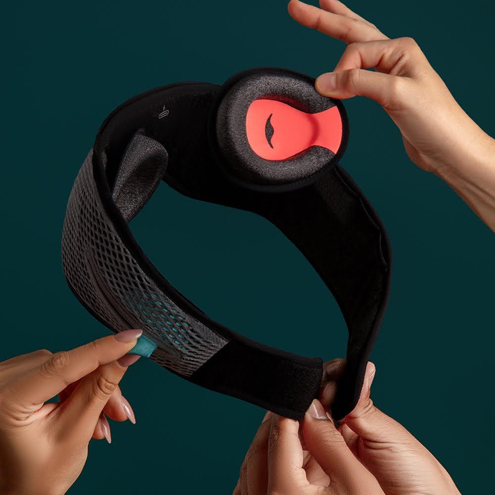Rear view of a gray sleep mask with headphones being held up by 3 hands. One hand is holding a C-shaped eye cup, the other is holding the blue pull tab of a speaker and a pair of hands is holding the micro hook-and-loop closure.