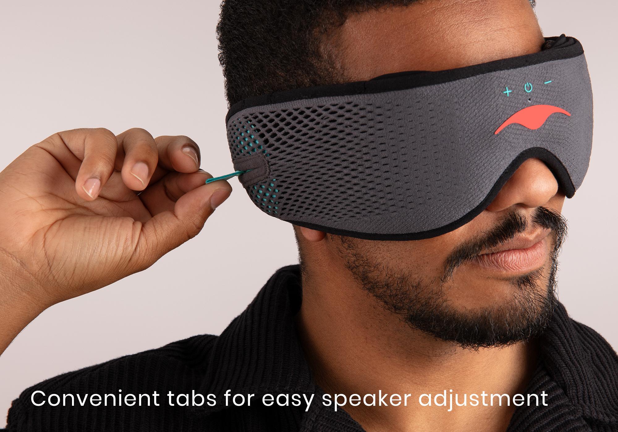A bearded man wearing a Bluetooth sleep mask with headphones, tugging on the tabs of adjustable speakers.