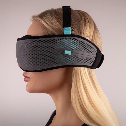 Side view of a blonde girl wearing a gray mesh sleep mask with headphones and a blue adjustable speaker. She is wearing a top strap for a more secure fit.